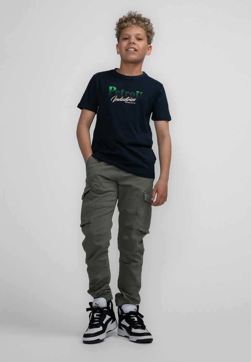 Marine t-shirt met groen en witte "Petrol Industries" grafiek, gecombineerd met olijfgroene cargobroek en zwart-witte high-top sneakers.