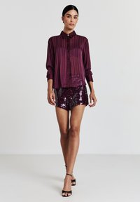 Gestreiftes Satin-Hemd in Kastanienbraun mit Knopfleiste, kombiniert mit glänzenden Pailletten-Shorts, die einen taillierten Schnitt und kurze Ärmel haben. Schwarze Riemchensandalen runden das Outfit ab.