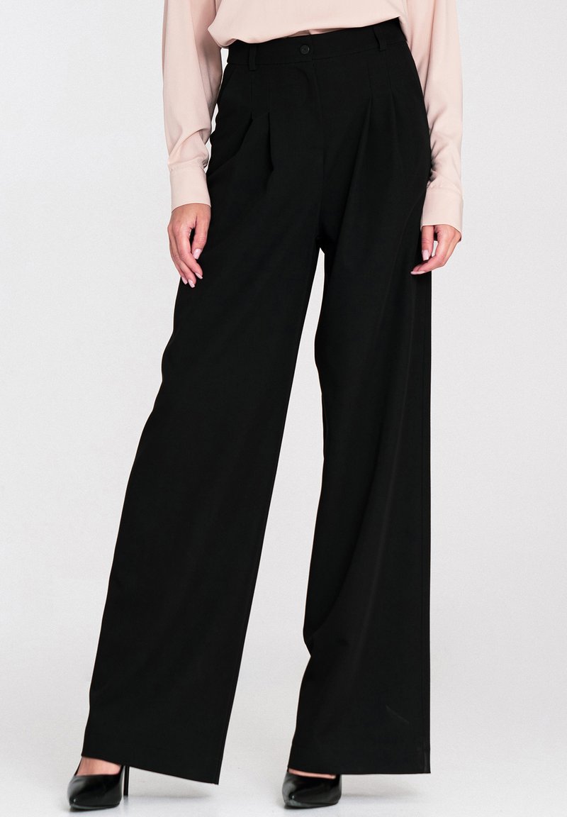 Czarne spodnie typu wide-leg z wysokim stanem, z fałdami z przodu i gładką teksturą. Połączone z jasnoróżową bluzką z długim rękawem.