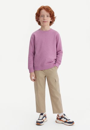 Niño con cabello rojo vistiendo una sudadera rosa, pantalones cargo beige y zapatillas blancas, azul marino y naranjas, de pie con una mano en el bolsillo.