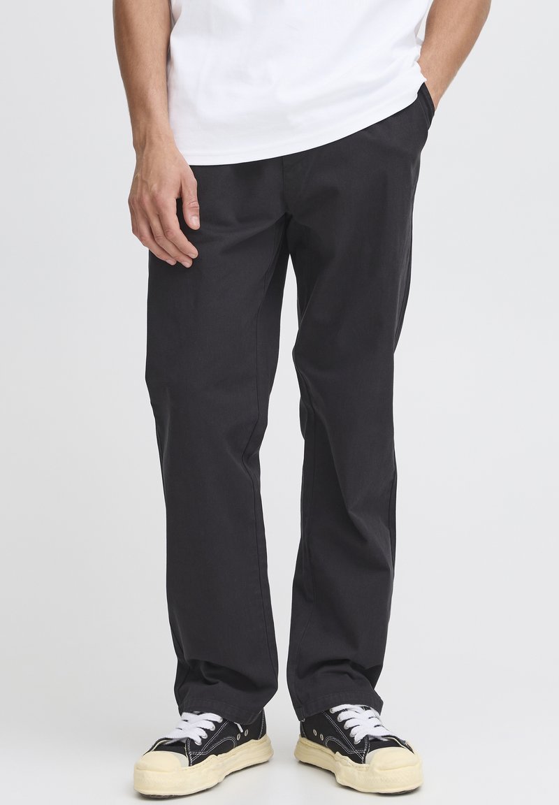 Black cotton pants with straight-cut legs, side pockets, and a smooth texture, worn with black sneakers featuring white laces and cream soles.