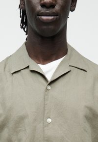 Lähivõte inimesest, kes kannab oliivirohelist nööbitavat särki valge t-särgi kohal, näidates alumist näopiirkonda, kaela ja ülemist rinnaosa.