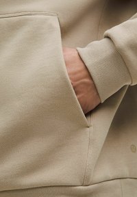 Beige-coloured collegepaita, jossa on ribbestatut hihansuut; varustettu sivutaskulla. Materiaali on sileä ja siinä on vähäisesti sauman yksityiskohtia.