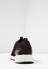 Vinröd träningssko med överdel i andningsbar stickad textil, texturerade detaljer, dragflik och vit vadderad mellansula med svart yttersula.