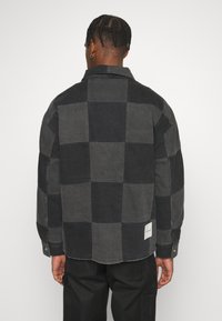 Schwarz- und graukariertes Hemd mit Kragen, langen Ärmeln und einem Etikett unten links. Der Stoff hat eine strukturierte Oberfläche.