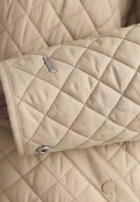 Bézs színű steppelt anyag gyémánt alakú varrással, fém patenttal és egy kis téglalap alakú márkajeggyel az ujj mandzsettáján egy ruhadarabon.