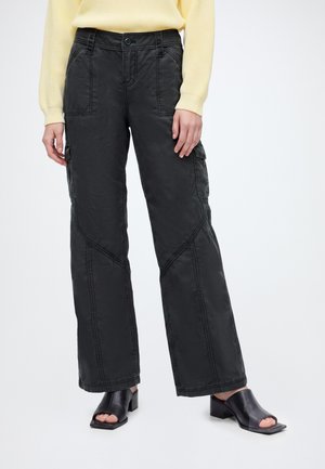 Personne portant un pantalon cargo noir ample, un haut jaune à manches longues et des mules à talons noires ouvertes, debout sur un fond blanc.