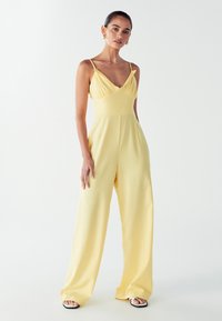 Žlutý overal s širokými nohavicemi, tenkými ramínky a výstřihem do V. Tkanina vypadá lehce a má hladkou strukturu. Černé sandály doplňují outfit.