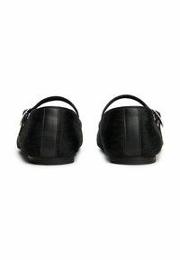 Par de zapatos planos negros con correas al tobillo, textura de piel de animal y una tira de cuero en la parte trasera, mostrados desde atrás sobre un fondo blanco.