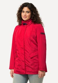 Giacca rossa a maniche lunghe con colletto alto, chiusura con zip e tasche laterali. Presenta un tessuto liscio e leggero e una toppa con logo sulla manica.