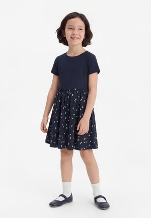 Uśmiechnięta dziewczyna z krótkimi ciemnymi włosami, ubrana w granatową sukienkę z kwiecistą spódnicą, białe skarpetki i granatowe buty Mary Jane.