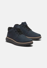 Zapatos de ante azul oscuro hasta el tobillo con cordones negros y un collar acolchado negro. Presentan un pespunte en contraste marrón sobre una suela negra.