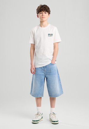 Weißes Baumwoll-T-Shirt mit grünem "Good Life"-Druck, kombiniert mit hellblauen Jeansshorts. Das Modell trägt weiße Sneaker und weiße Socken.