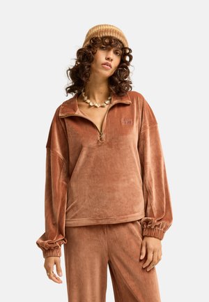 Frau mit lockigem Haar trägt ein braunes Cord-Reißverschluss-Top und passende Hose, eine beige Strickmütze und eine auffällige Muschelkette.