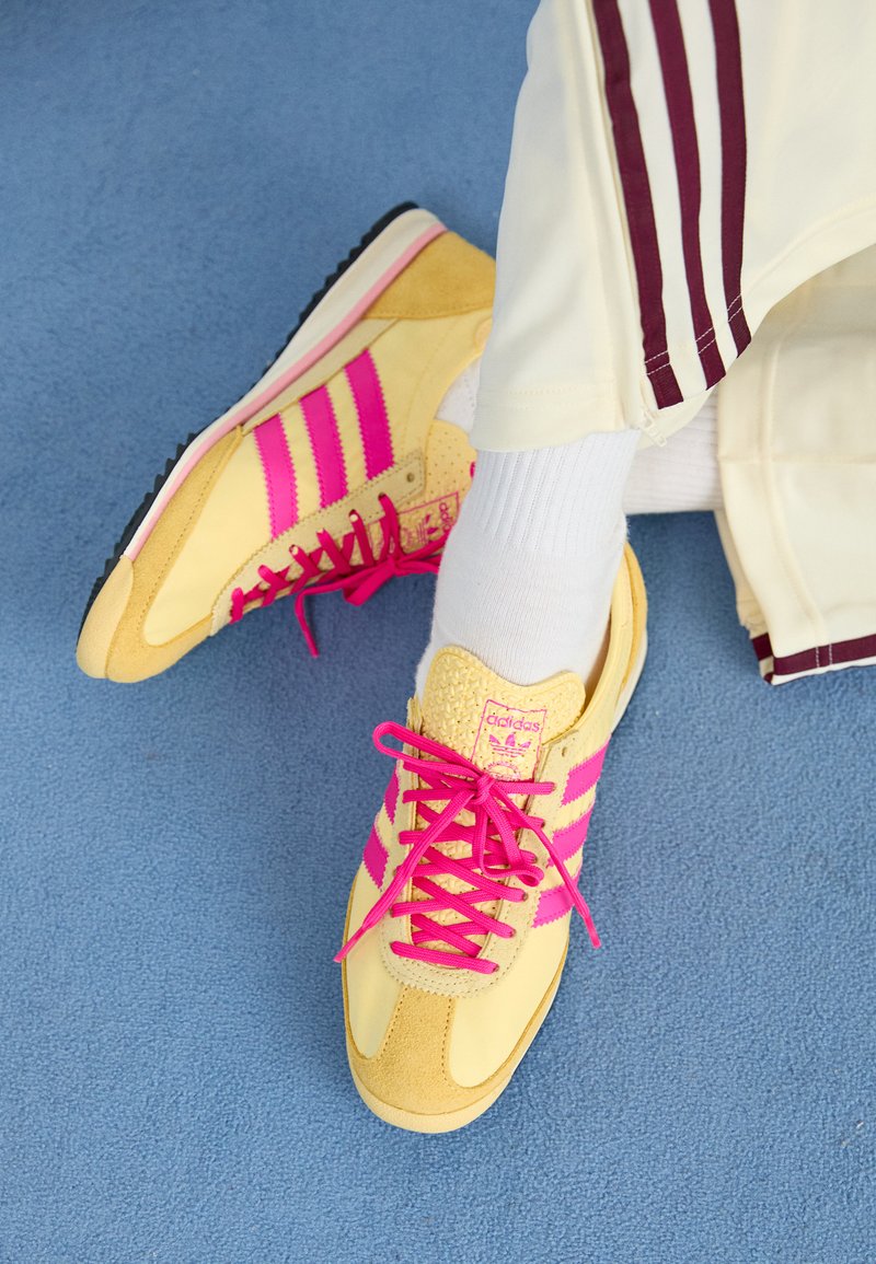 Gelbe Sneakers mit pinken Akzenten, mit einem Design aus Wildleder und Mesh, auffälligen Schnürsenkeln und Streifen, vor einem blauen Hintergrund.