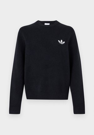 Młody mężczyzna z kręconymi włosami, ubrany w czarny sweter z okrągłym dekoltem z białym logo Adidas oraz ciemne sprane jeansy, stoi na tle jednolitego tła.