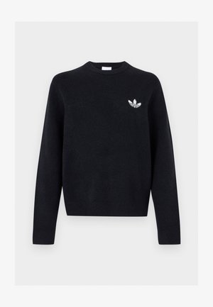 Młody mężczyzna z kręconymi włosami, ubrany w czarny sweter z okrągłym dekoltem z białym logo Adidas oraz ciemne sprane jeansy, stoi na tle jednolitego tła.