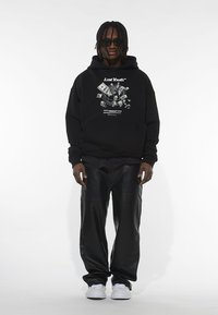 Crna dukserica s grafičkim printom, u paru s labavim crnim kožnim hlačama i bijelim tenisicama. Model nosi sunčane naočale i ima pletenu kosu.