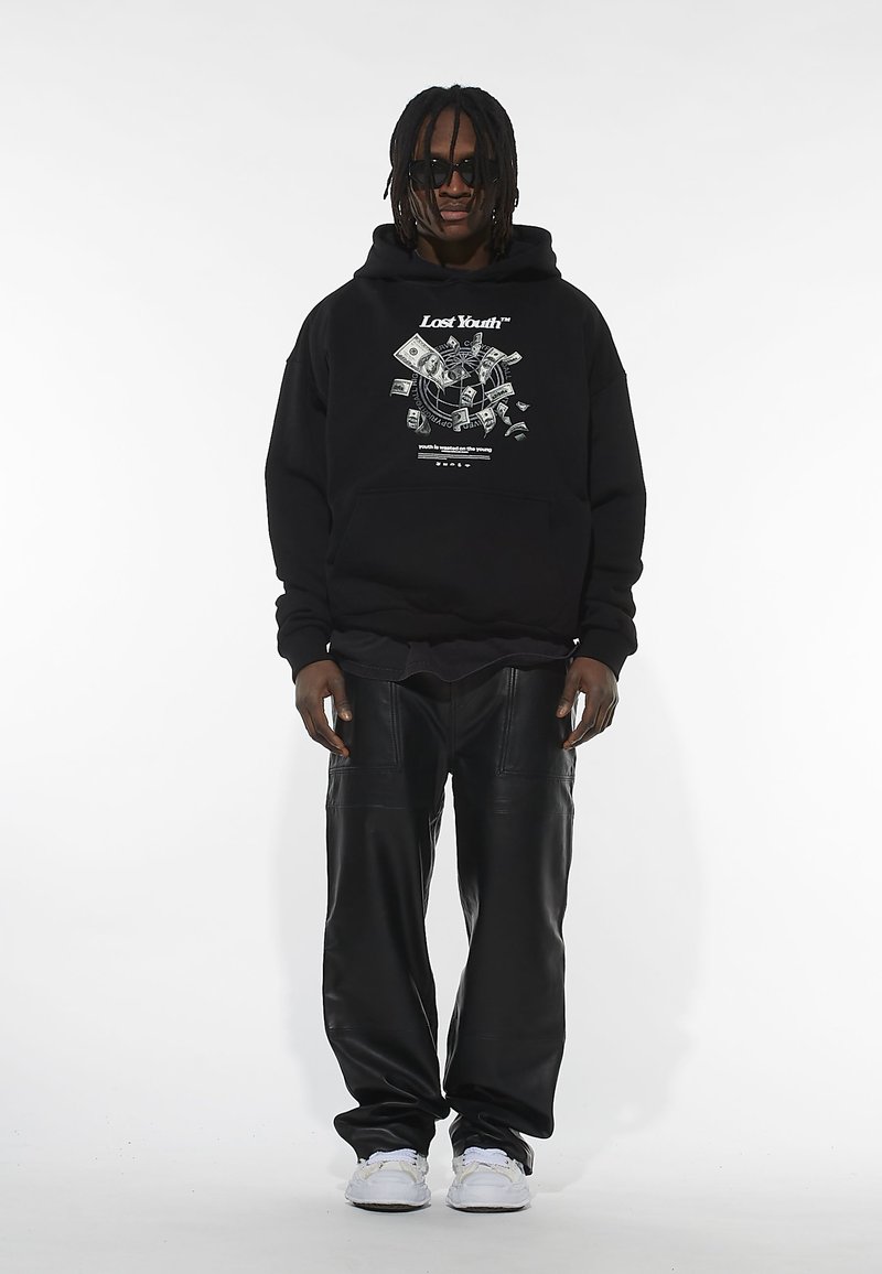 Crna dukserica s grafičkim printom, u paru s labavim crnim kožnim hlačama i bijelim tenisicama. Model nosi sunčane naočale i ima pletenu kosu.