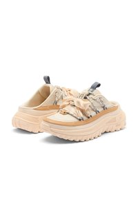 Chunky beige slip-on sneakers with thick rubber soles, crisscross laces, a large fabric bow on the front, and pull tabs on the tongues.