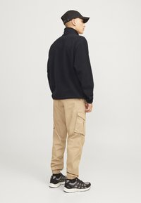 Pull en polaire noir avec col montant, pantalon cargo beige avec poches latérales, et baskets noires avec accents gris, vu de derrière.