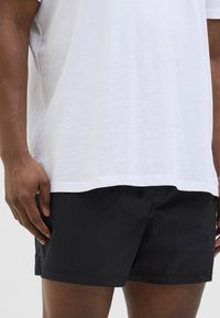 Shorts de bain noirs en tissu léger, présentant un design simple et une taille élastique. Portés avec une chemise blanche ample.