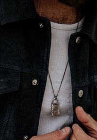 Silverlås-hängsmycke på en kedja hänger mot en ribbad vit skjorta, delvis dold av en svart jacka med metallknappar.