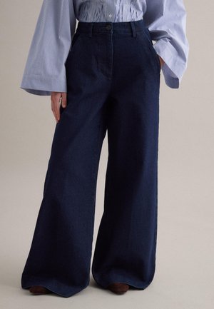 Donkere denim broek met wijde pijpen, gedragen met een ingestopte blauw-wit gestreepte blouse met wijde mouwen, handen in de zakken.
