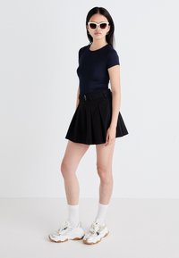 Marineblaues, tailliertes T-Shirt, schwarzer Faltenrock mit Gürtel, weiße Crew-Socken, silberne Turnschuhe mit Gummisohlen und weiße übergroße Sonnenbrille.