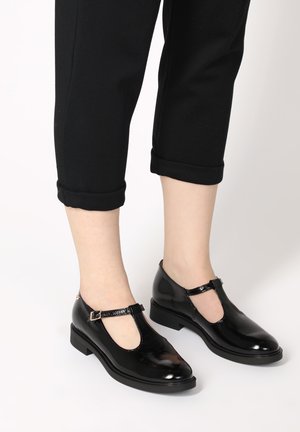 Black patent leather T-strap shoes with a rounded toe, low block heel, and adjustable ankle strap. Smooth finish and minimal detailing.