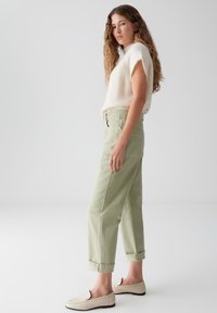Haut léger en tricot beige crémeux associé à un pantalon léger vert clair à taille haute et coupe décontractée. Le modèle porte des chaussures beiges à enfiler.