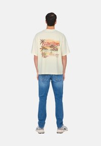 Cremefarvet bomulds oversized t-shirt med et farverigt palmetræ grafisk design og tekst på ryggen, parret med lyseblå denim jeans og sneakers.