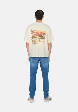 Cremfarbener, übergroßer Baumwoll-T-Shirt mit einem bunten Palma-Baum-Graphic und Text auf der Rückseite, kombiniert mit hellblauen Jeans und Sneakers.