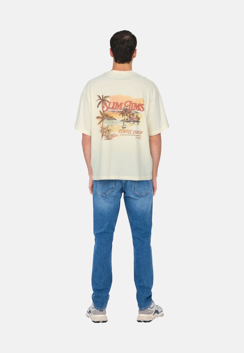 Cremefarvet bomulds oversized t-shirt med et farverigt palmetræ grafisk design og tekst på ryggen, parret med lyseblå denim jeans og sneakers.