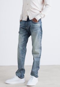 Gai zila džinsu bikses ar brīvu piegriezumu, ar izbalējuma detaļām, izbalējušu krāsu un piecu kabatu dizainu, kombinētas ar baltām sporta apaviem.