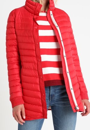 Rød polstret jakke med høj krave, trykknapper og sidelommer. Båret over en rød og hvid stribet sweater, parret med blå jeans.