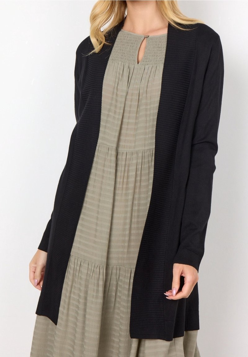 Femme portant une robe plissée beige avec un col en forme de goutte et un long cardigan noir ouvert par-dessus.