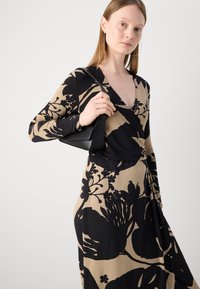 Musta ja beige kukkakuosiset kietaisumekko pitkillä hihoilla, jossa on syvä V-pääntie ja hienovarainen tekstuuri. Mukana pieni musta käsilaukku.