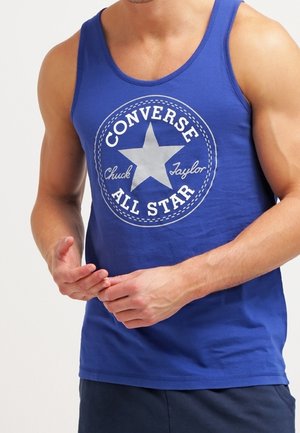Mężczyzna w niebieskim bezrękawniku z białym logo Converse All Star i granatowych szortach, ręce złożone z przodu.