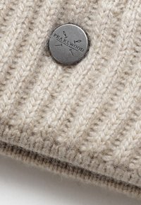 Beige pletená čepice s vertikálním žebrováním, s kulatou kovovou známkou s vyraženým nápisem "PEARLWOOD". Měkký, teplý vlněný materiál.