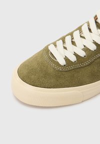 Oļīvu zaļas zīda zamšāda sporta kurpes ar baltām aukliņām, apaļu purngalu un krēmkrāsas gumijas zolīti ar teksturētām detaļām pa malām.