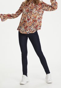 Mujer posando con jeans ajustados oscuros, zapatillas deportivas blancas y una blusa floral de manga larga con puños acampanados, sobre un fondo blanco liso.