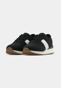 Μαύρα αθλητικά παπούτσια με λευκή ρίγα, διαθέτουν ανθεκτικό υλικό άνω μέρους, επενδυμένο γιακά και ελαφριά καφέ λαστιχένια σόλα με υφή για καλύτερο κράτημα.