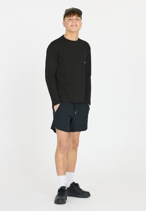 Svart långärmad skjorta, mörka shorts, vita strumpor och svarta sneakers. Modellen står med händerna i fickorna mot en enkel bakgrund.