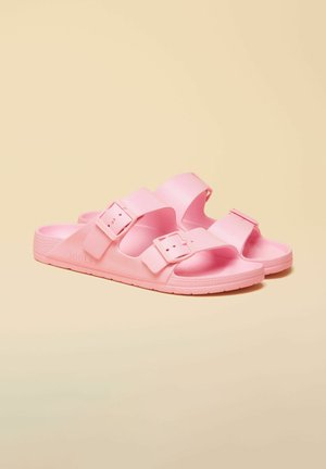 Pár ružových šmykľavých sandálov s dvoma nastaviteľnými prackami na béžovom pozadí.