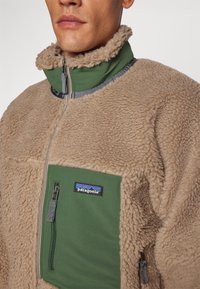 Beige fleece jacket with green fabric panel. Features zip front, high collar, and zip pocket. Soft texture, minimal hardware accents.