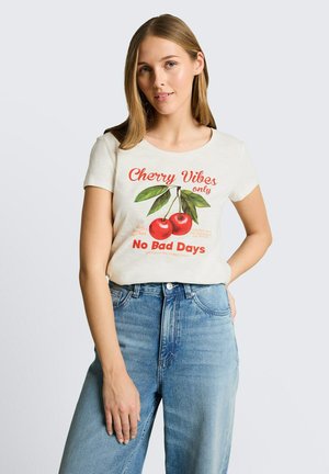 Kobieta ubrana w białą koszulkę z czerwonymi wiśniami i napisem „Cherry Vibes only No Bad Days”, w połączeniu z niebieskimi, wysokim stanem jeansami, na jednolitym tle.