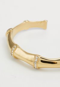 Bracciale a bangle in tonalità oro a forma di bambù, con superficie liscia e dettagli in cristallo trasparente. Presenta bordi arrotondati e un'apertura flessibile.