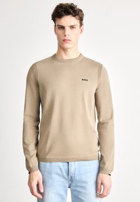 Beżowy sweter z dzianiny z długimi rękawami, okrągłym dekoltem i subtelnym prążkowanym zdobieniem na ramionach. Posiada małe czarne logo "BOSS".