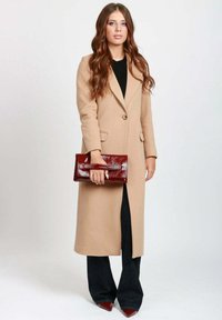 Donna con capelli lunghi castani, che indossa un lungo cappotto beige, pantaloni neri, scarpe a punta bordeaux e tiene in mano una brillante pochette bordeaux.
